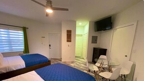 A hotel room with two beds, a ceiling fan, a TV, a small table with chairs, and a door leading to a hallway.
