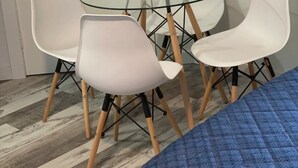 A room with a glass-top table surrounded by white chairs with wooden legs.
