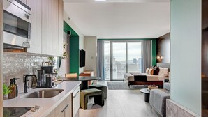 A modern kitchen with a built-in oven, a dining area with a round table, and a living room with a sofa and a large window offering a city view.