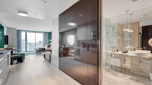 A modern hotel room with a large window, a kitchen area, and a bathroom with a marble countertop.