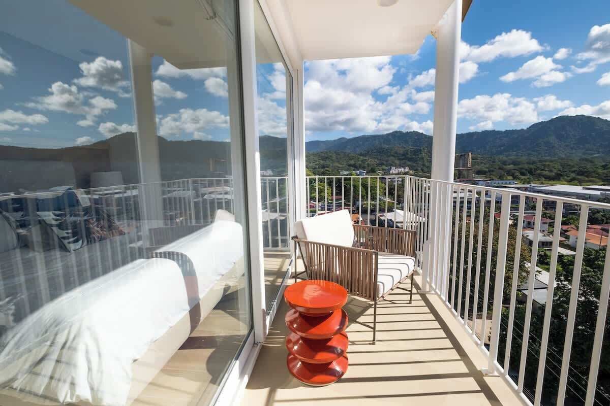 A balcony with a bed, a table, and a chair, overlooking a mountainous landscape.