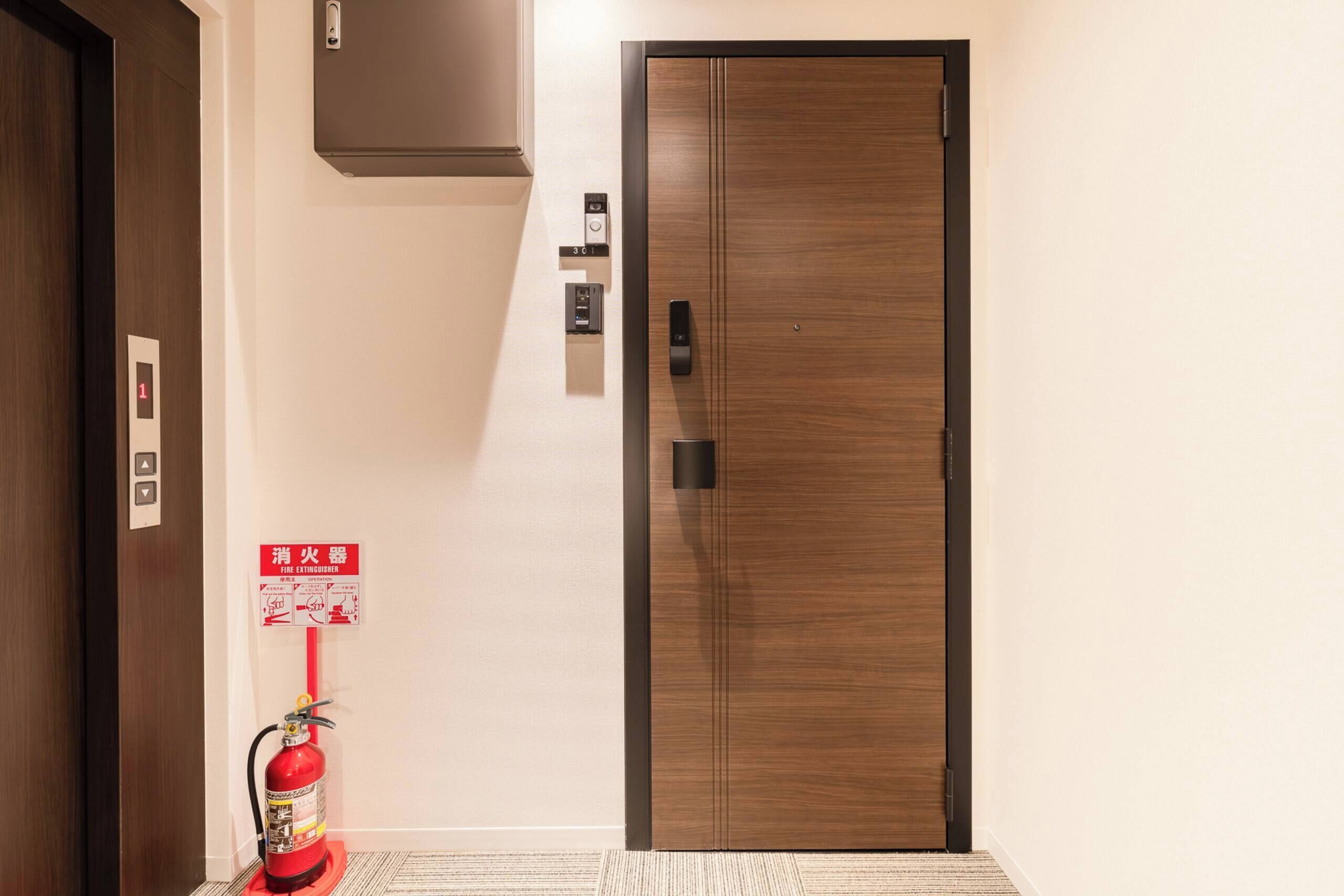 A hotel room entrance with a wooden door, a fire extinguisher, a fire safety sign, and a wall-mounted control panel.