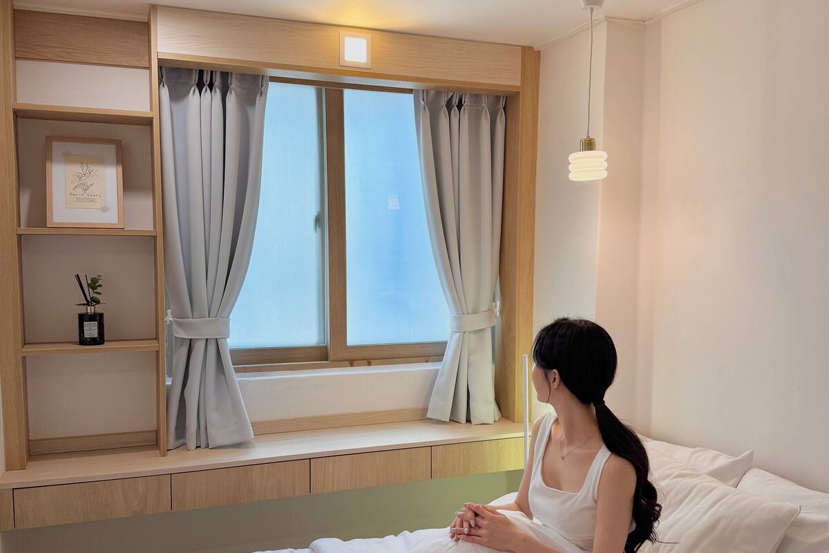 A room with a bed, a window with curtains, a shelf with a framed picture, and a bedside lamp.