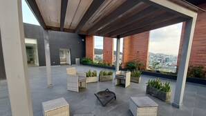 A rooftop terrace with wicker furniture, potted plants, and a view of the city.