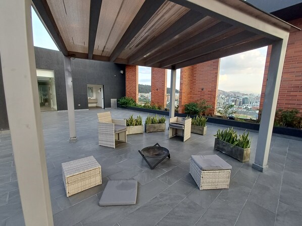 A rooftop terrace with wicker furniture, potted plants, and a view of the city.