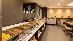 Un buffet proposant une variété de plats, un espace repas avec des tables et des chaises, et un mur décoratif agrémenté d’une sculpture d’arbre.