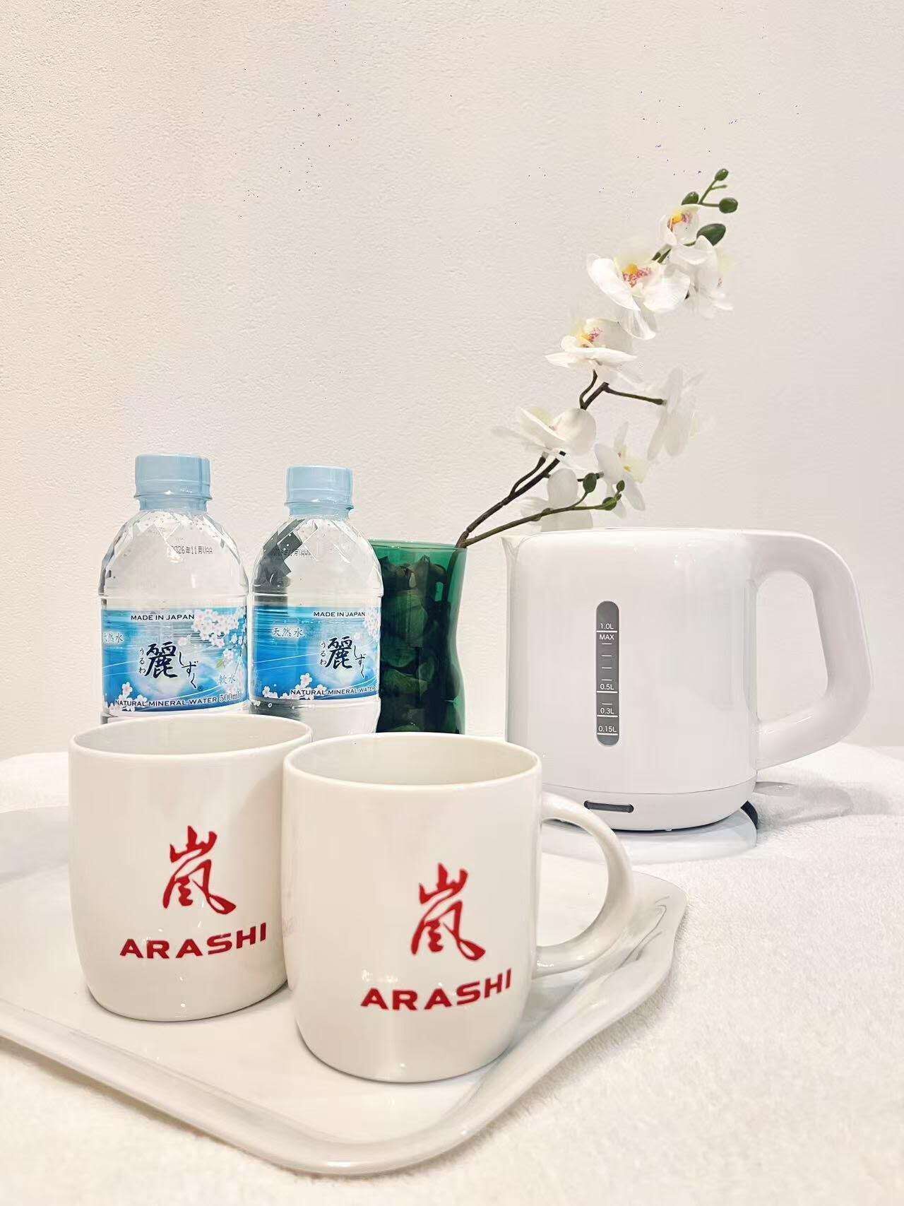 Deux bouteilles d’eau, deux tasses ornées du mot « ARASHI », un vase vert avec des fleurs et une bouilloire électrique blanche, le tout disposé sur un plateau.