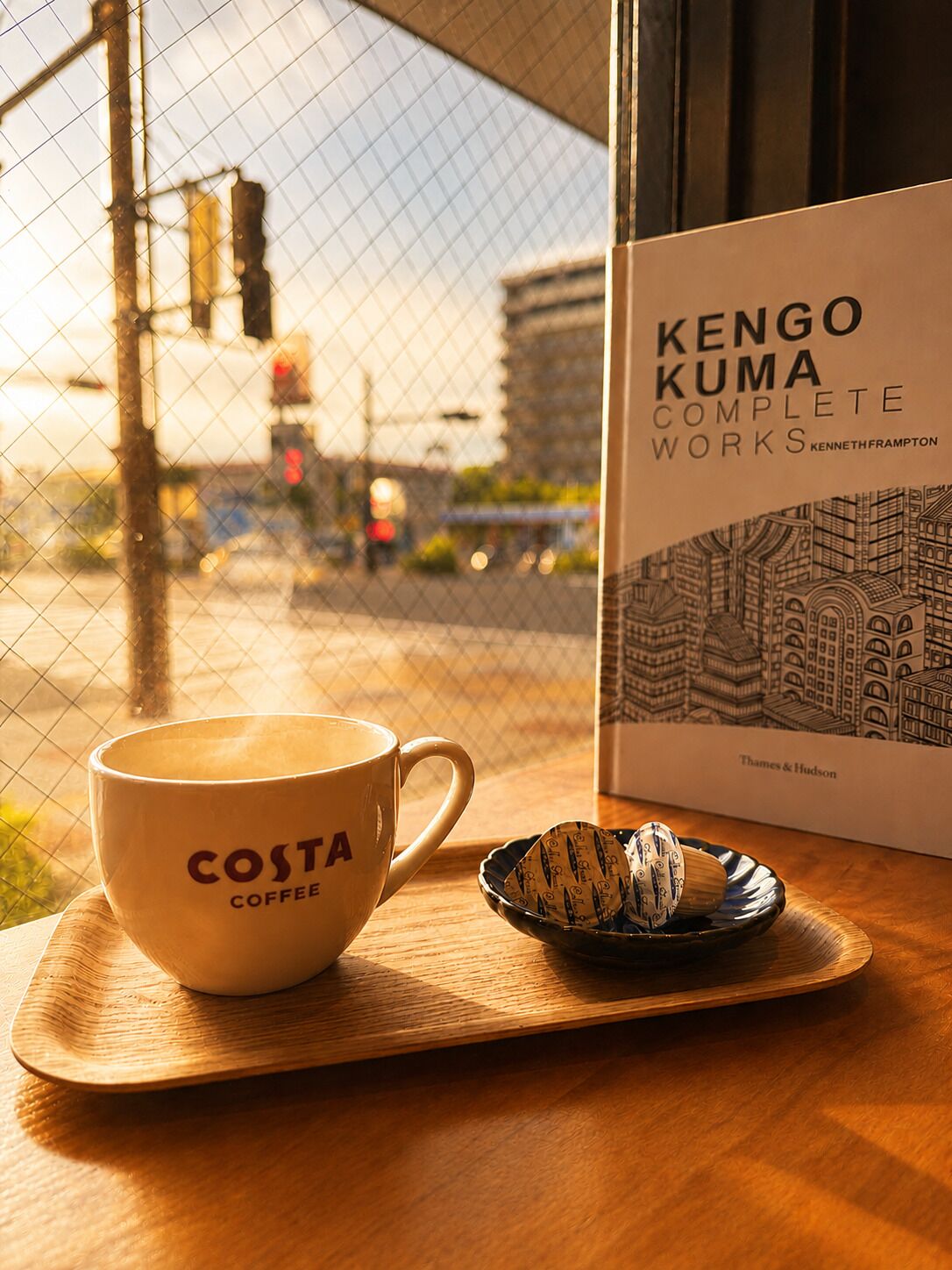 Een koffiekop met de tekst 'COSTA COFFEE', een houten dienblad, een boek getiteld 'KENGO KUMA COMPLETE WORKS' van Kenneth Frampton, en een uitzicht op een stadsgezicht door een raam.