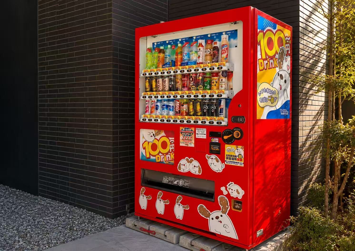 A red vending machine with various drink options and stickers, located against a brick wall.