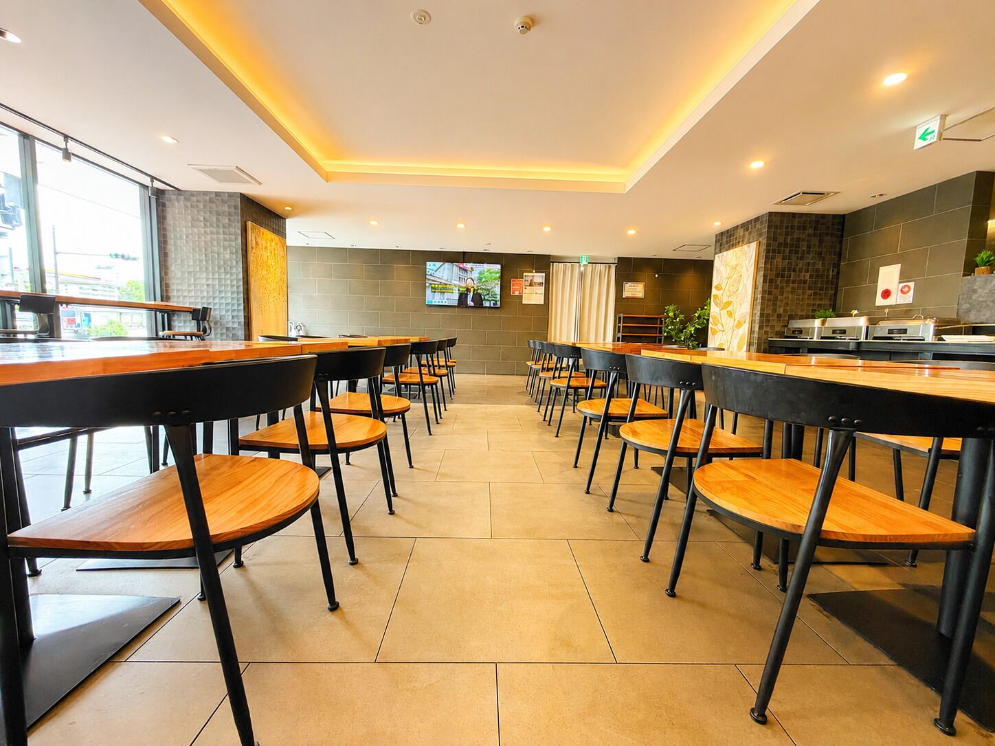 A modern dining area with long tables and chairs, a kitchen area, and a television screen.