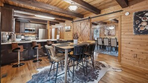 A rustic interior with a wooden ceiling, a bar area with stools, a dining table with chairs, and a kitchen in the background.