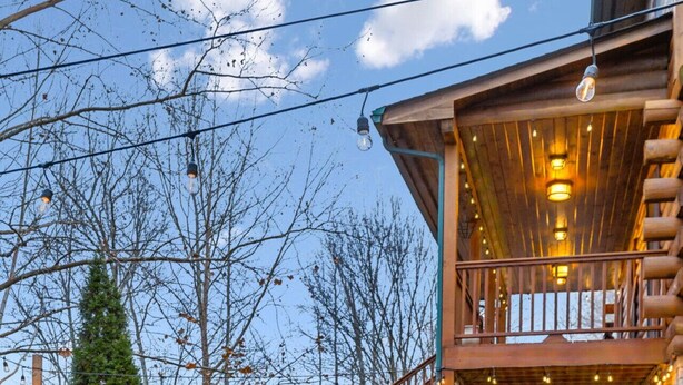 A cozy outdoor seating area with Adirondack chairs around a stone fire pit, a wooden deck with a railing, and a stone building with a balcony.
