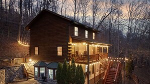 A two-story log cabin with a balcony, a stone staircase, and string lights.