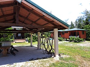 Un espace extérieur abrité avec une table et des chaises en bois, un petit bâtiment doté d’un porche et une zone herbeuse avec des arbres.
