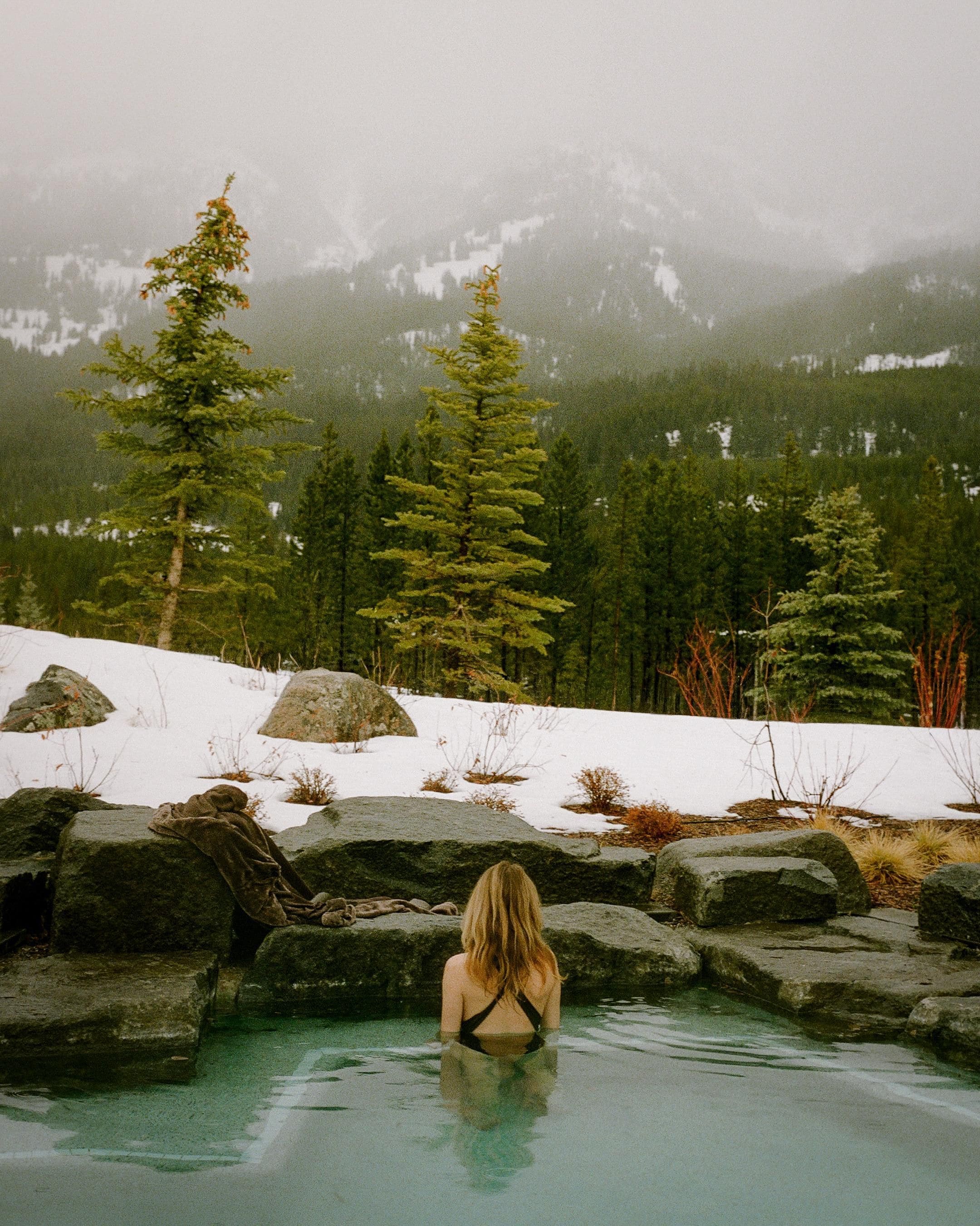 A person in a natural pool surrounded by rocks and snow, with trees and mountains in the background.