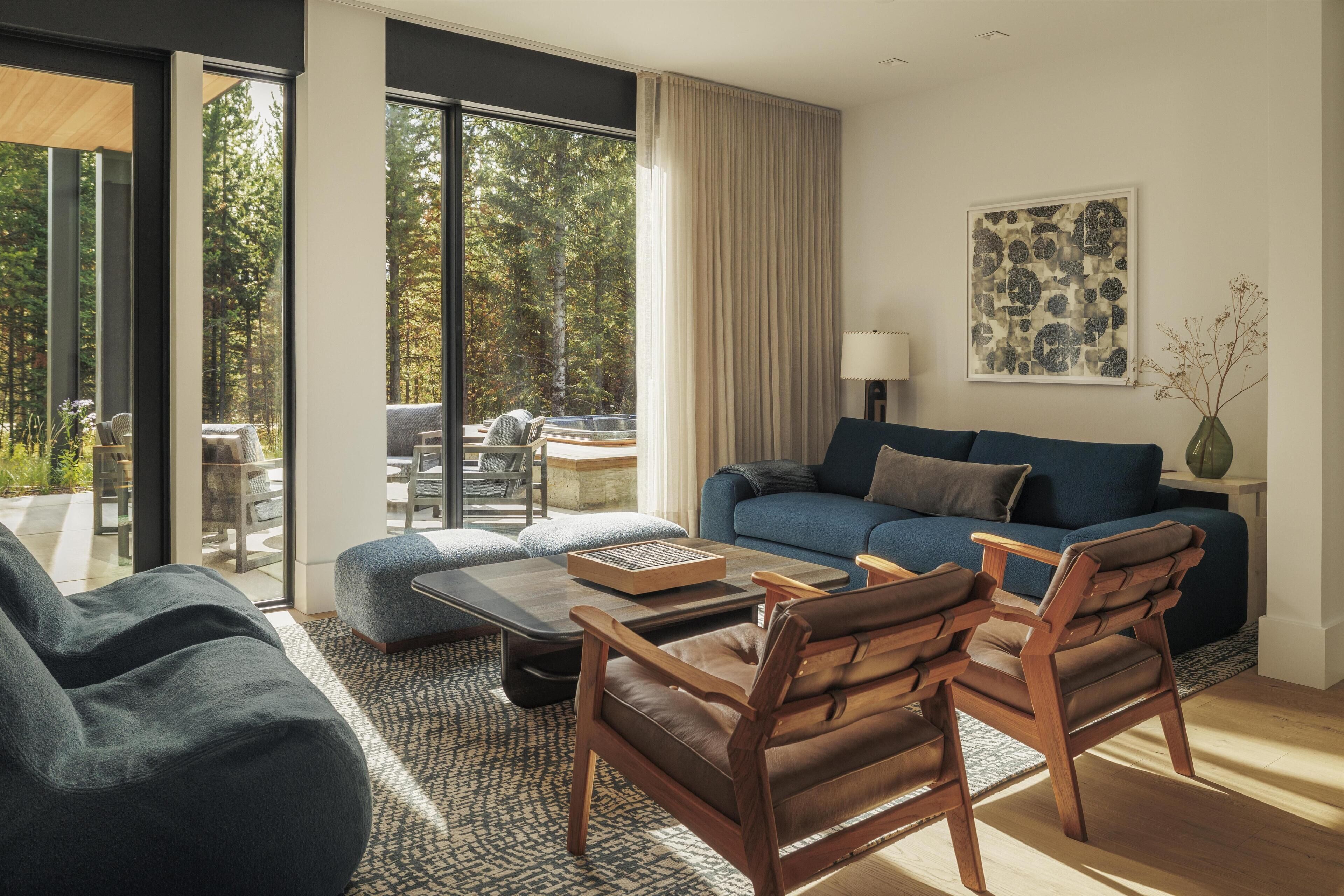 A modern living room with a blue sofa, wooden chairs, and a coffee table. Large windows offer a view of trees and a patio area.