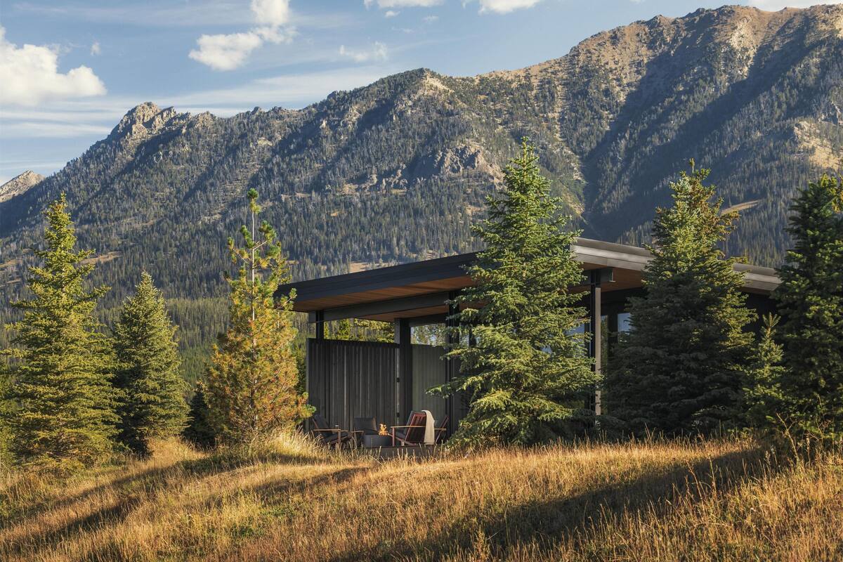 A modern cabin with a wooden exterior and glass walls, surrounded by tall pine trees and mountains in the background.