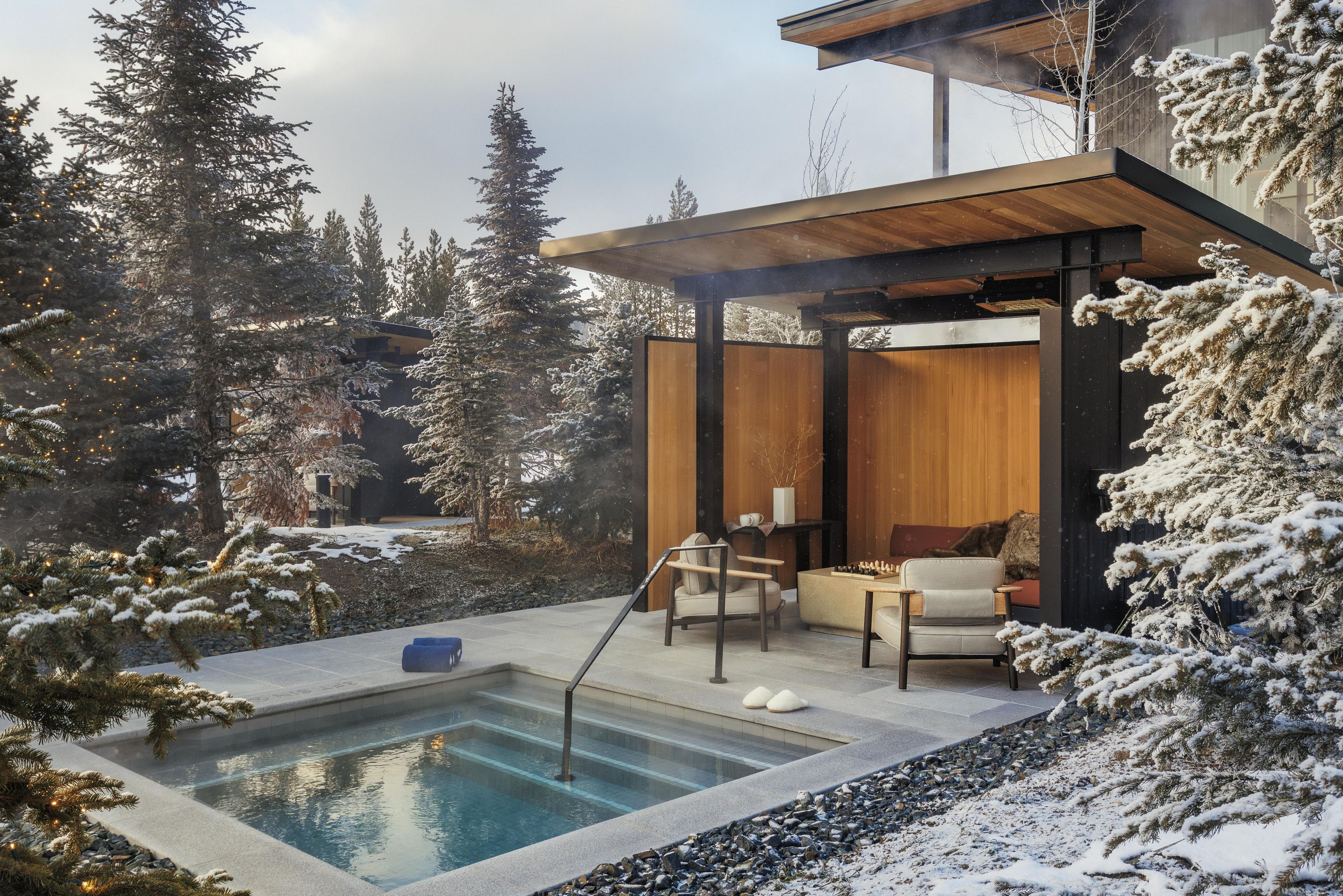 A modern outdoor pool area with a wooden deck, seating, and a view of snow-covered trees.