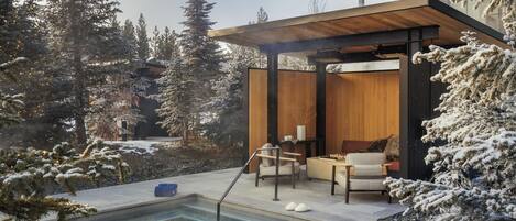 A modern outdoor pool area with a wooden deck, seating, and a view of snow-covered trees.