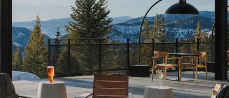 A cozy outdoor seating area with a fire pit, wooden chairs, and a table, overlooking a snowy mountain landscape.