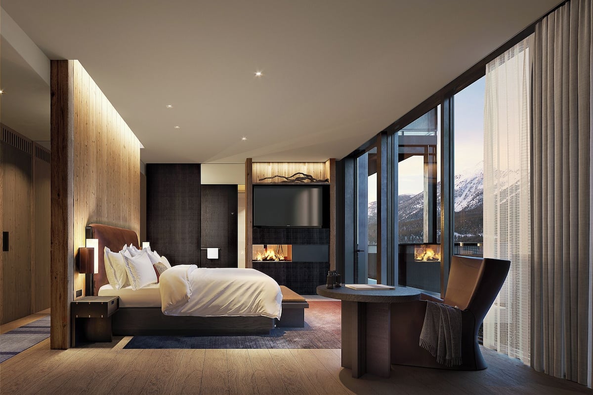 Una habitación de hotel moderna con una cama grande, una chimenea, un televisor y vistas a montañas a través de amplios ventanales.