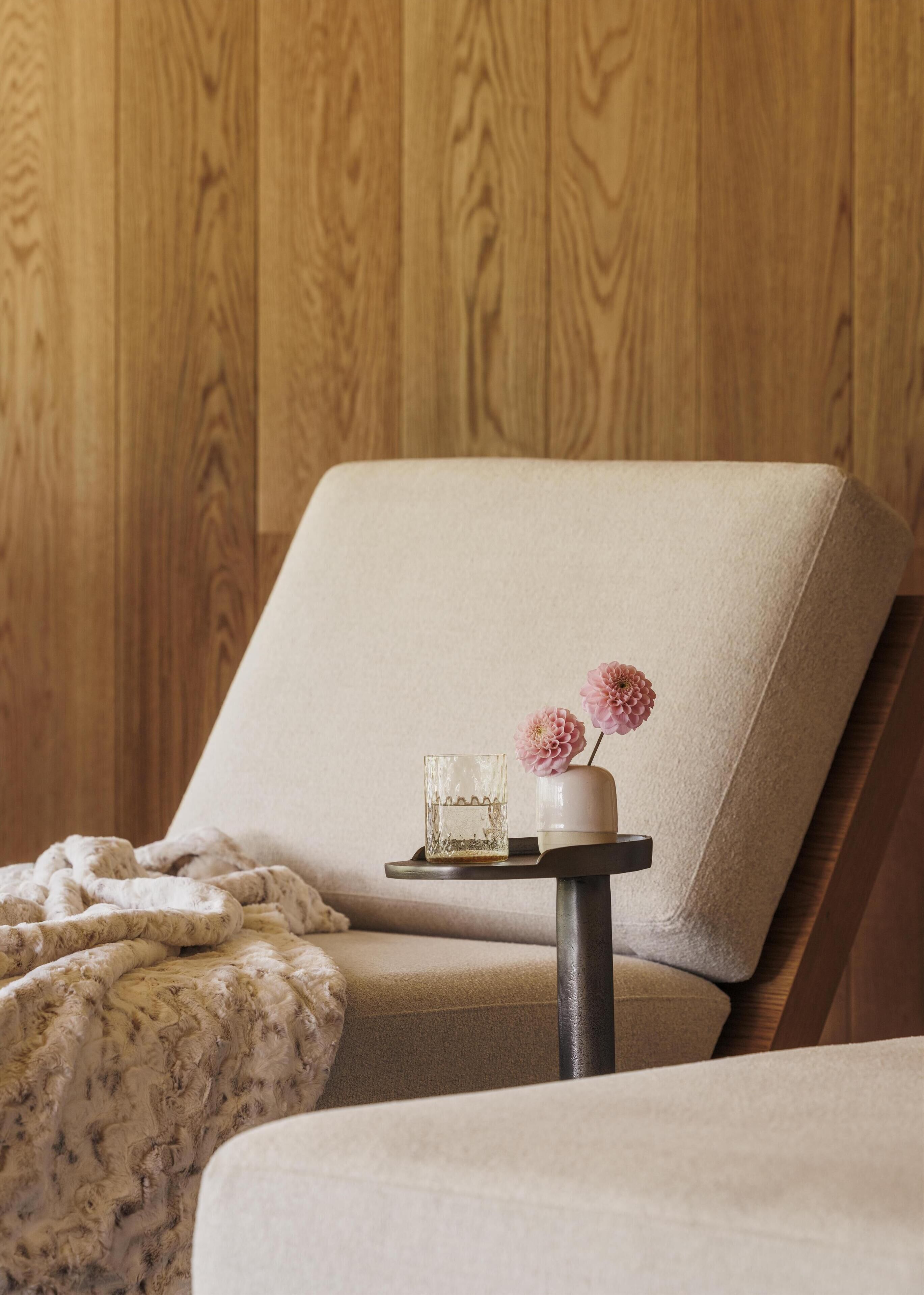 Una habitación acogedora con pared de madera, un sillón mullido, una mesita con un vaso y flores, y una manta estampada.