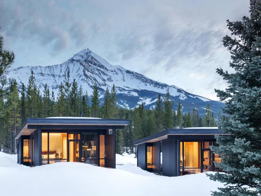 Two modern cabins with glass walls, surrounded by snow and trees, with a mountain in the background.