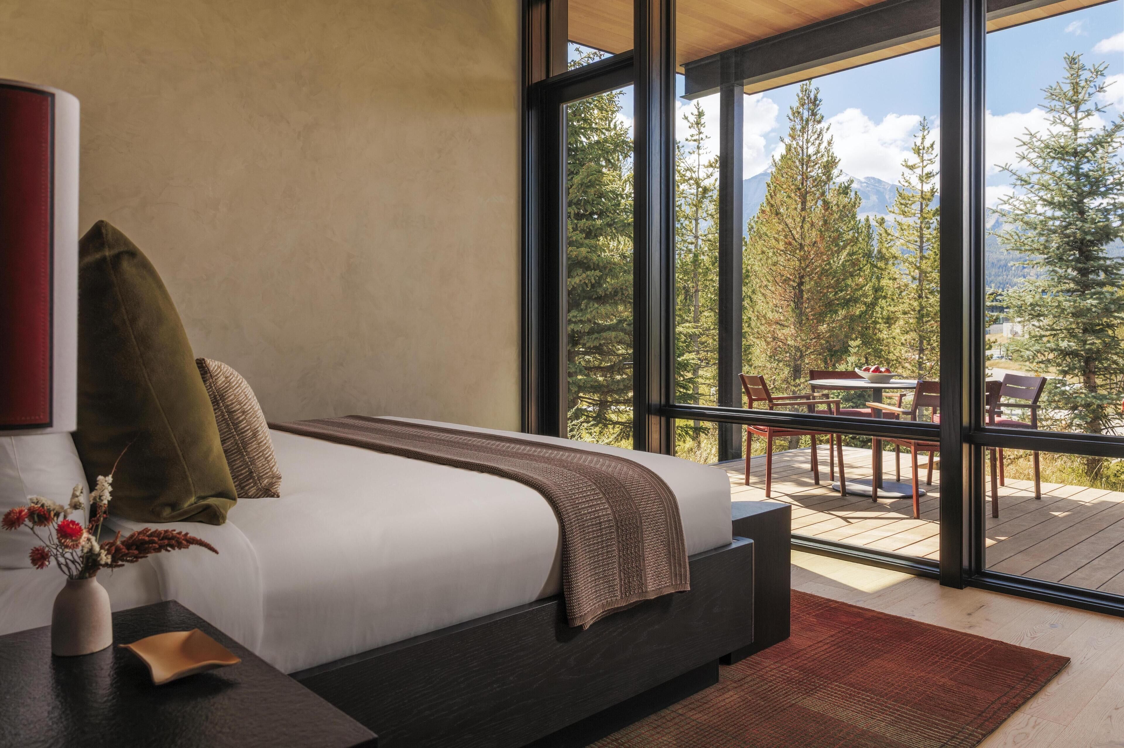 Un dormitorio moderno con una cama grande, vistas a una terraza con mobiliario exterior y montañas al fondo.