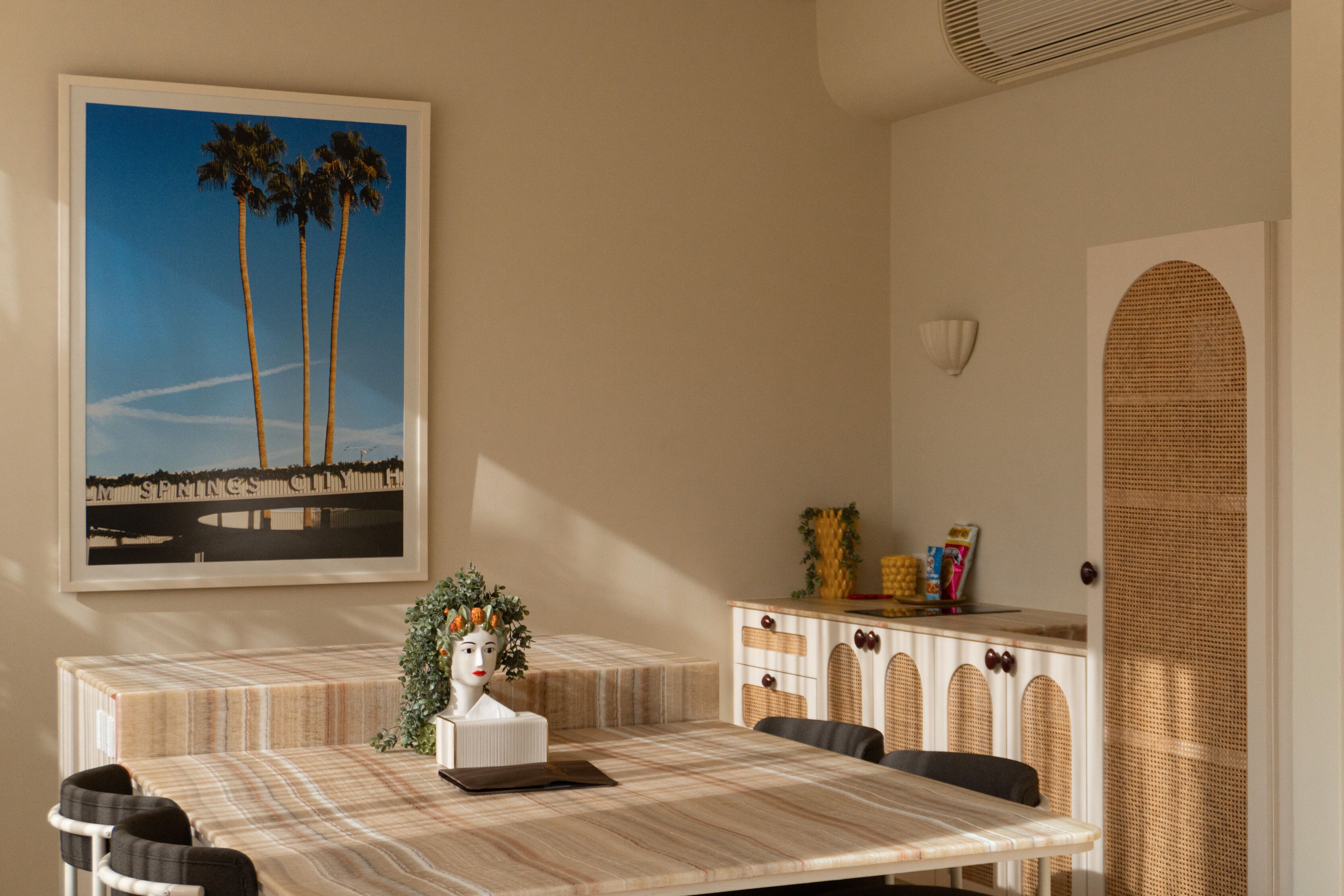 Un coin repas avec une table en marbre, des chaises, un tableau encadré représentant des palmiers, un buste décoratif et un meuble contenant un panier en osier.