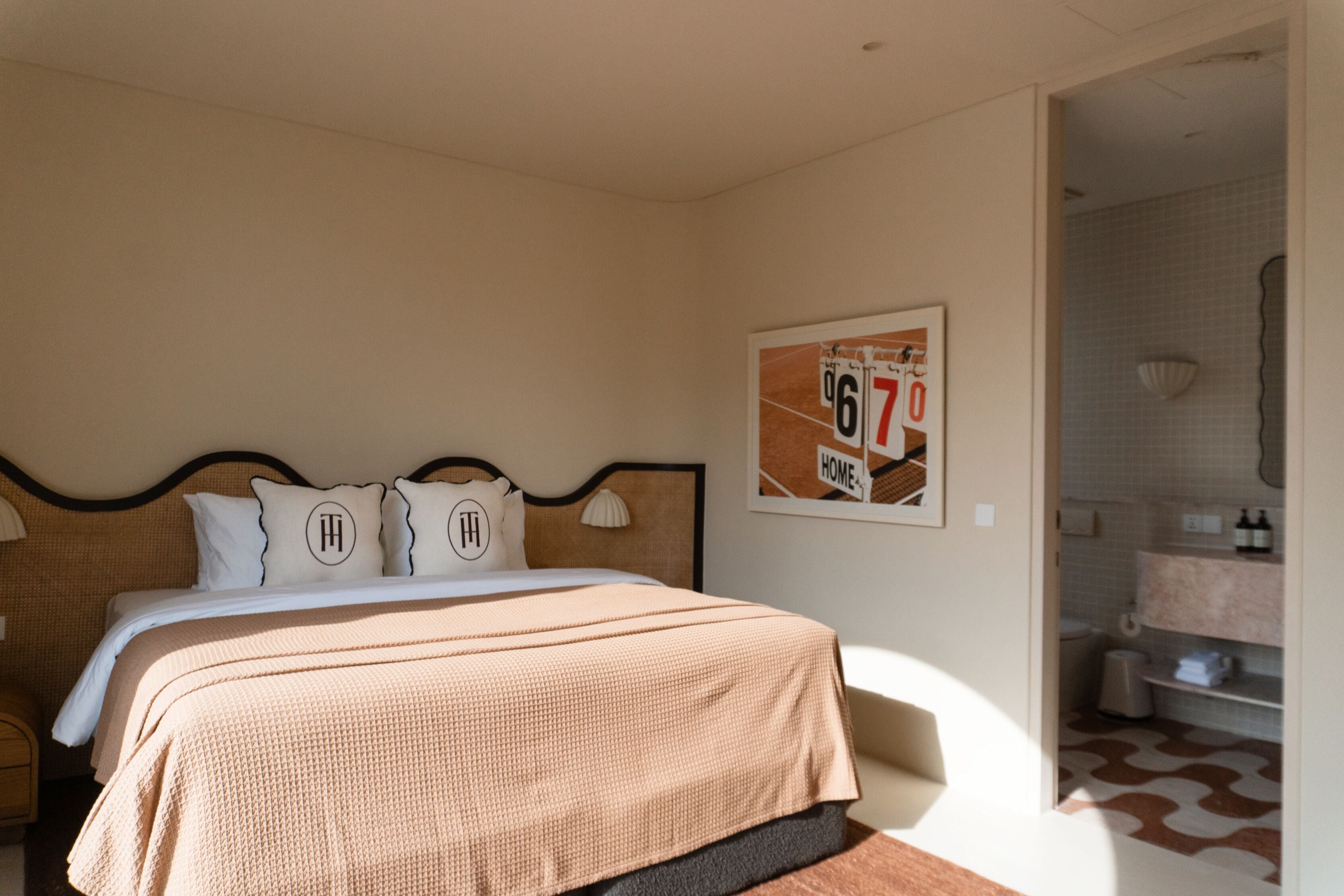 Une chambre à coucher avec un grand lit, une tête de lit, deux oreillers ornés des initiales brodées, un tableau encadré au mur et une salle de bain visible à travers une porte ouverte.