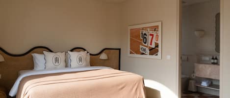 Un dormitorio con una cama grande, un cabecero, dos almohadas con iniciales bordadas, un cuadro en la pared y un baño visible a través de una puerta abierta.