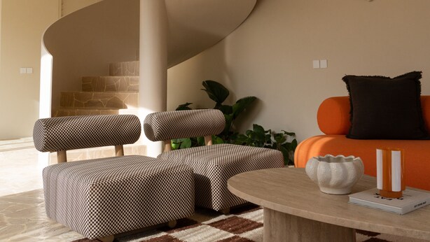 Un salon moderne avec un escalier à volutes, un canapé agrémenté d’un coussin noir, un pouf à motifs et une petite table sur laquelle sont posés un livre et une bougie.