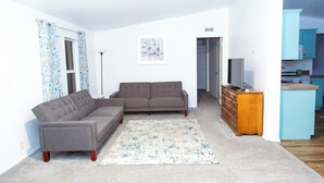 Un salón con un sofá seccional gris, un aparador de madera y una cocina visible a través de una puerta abierta.