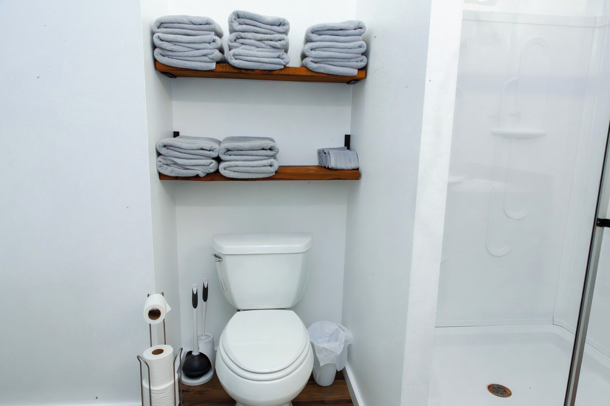 A bathroom with a toilet, two shelves with folded towels, a toilet paper holder, a plunger, and a shower area.