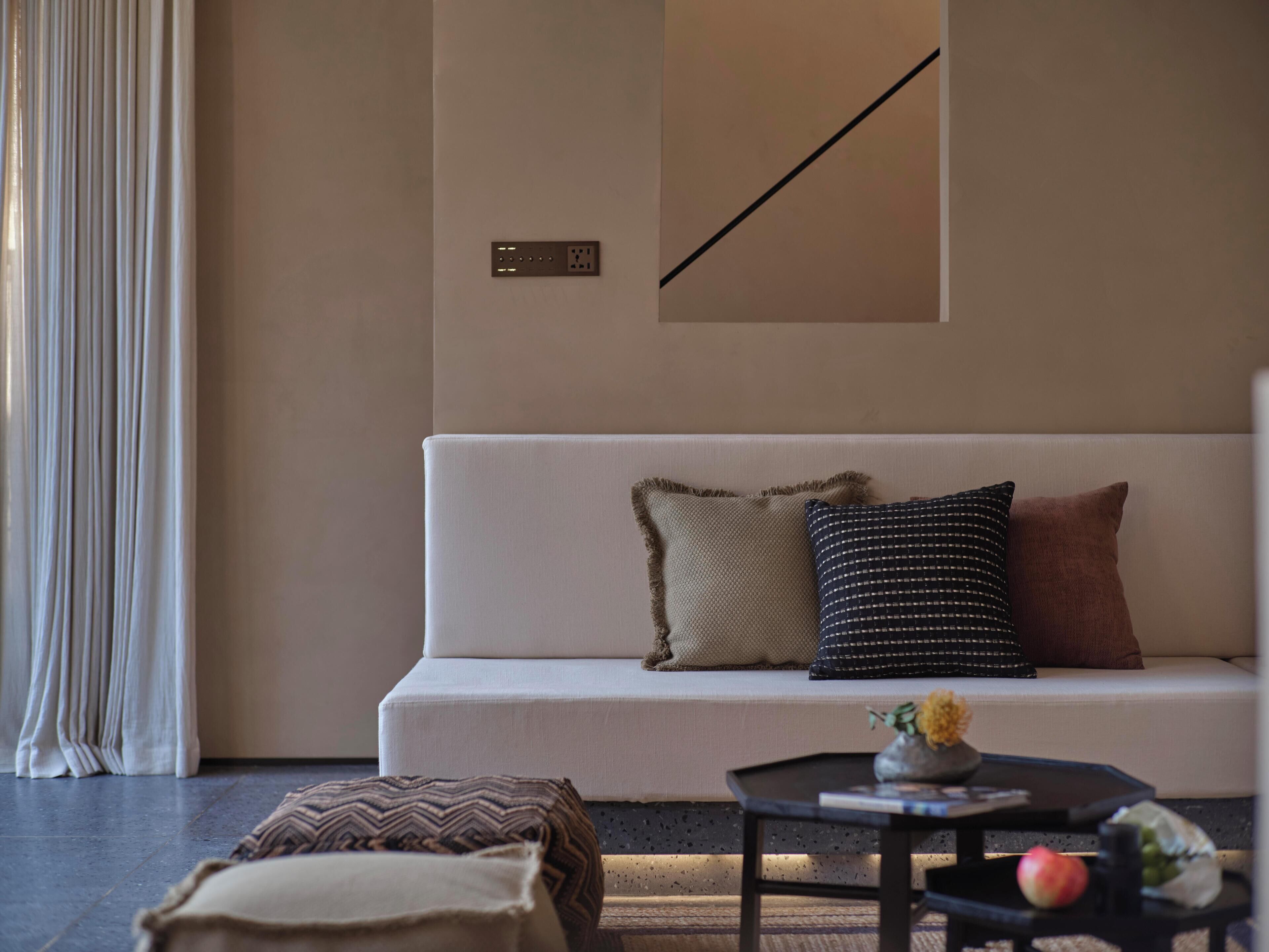 A modern living room with a white sofa, patterned cushions, a small side table with a vase and fruit, and a wall-mounted power outlet.