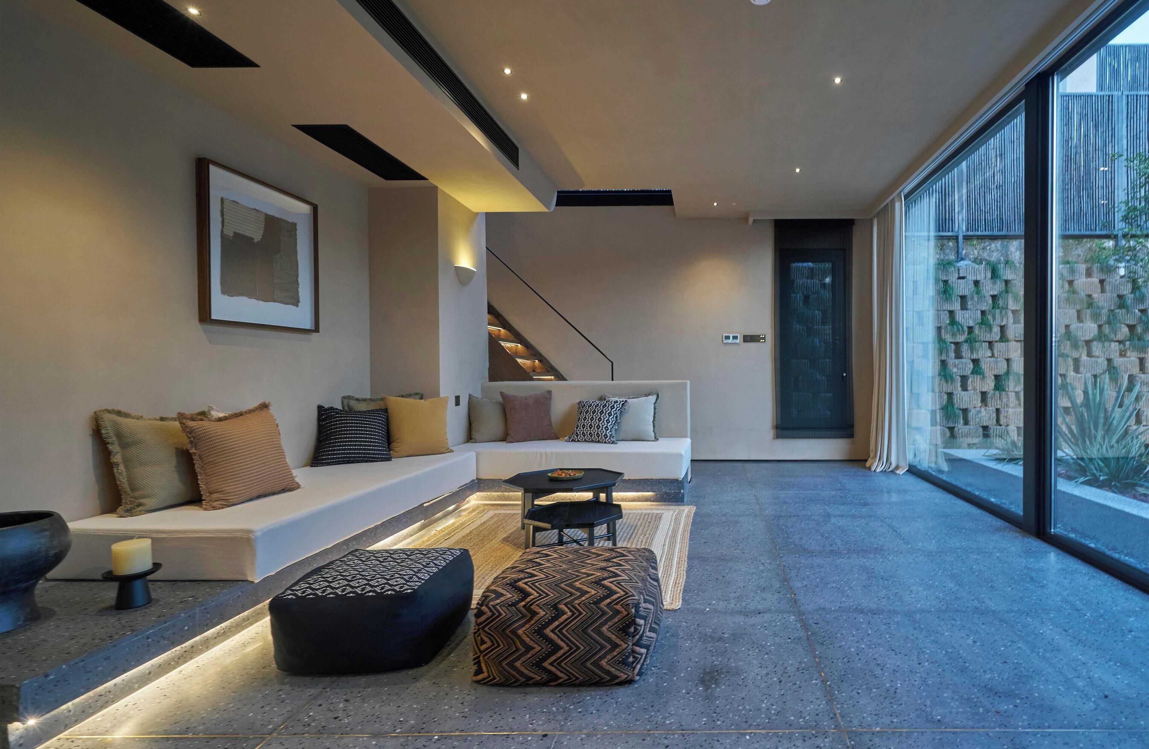 A modern living room with a white sofa, black ottoman, and a small table. There is a staircase leading to another level, and a large window with a view of greenery.