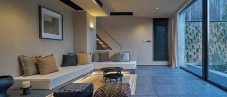 A modern living room with a white sofa, black ottoman, and a small table. There is a staircase leading to another level, and a large window with a view of greenery.