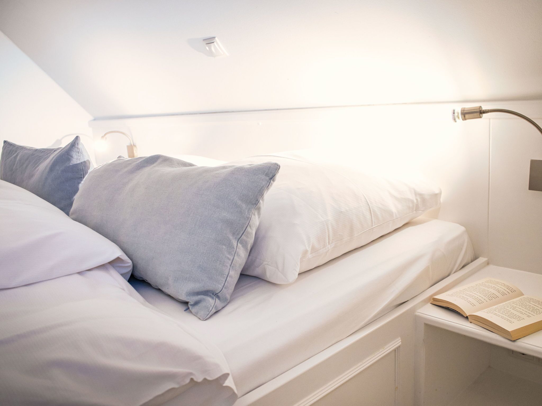 A neatly made bed with white and gray pillows, a bedside table with a book, and a modern light fixture.