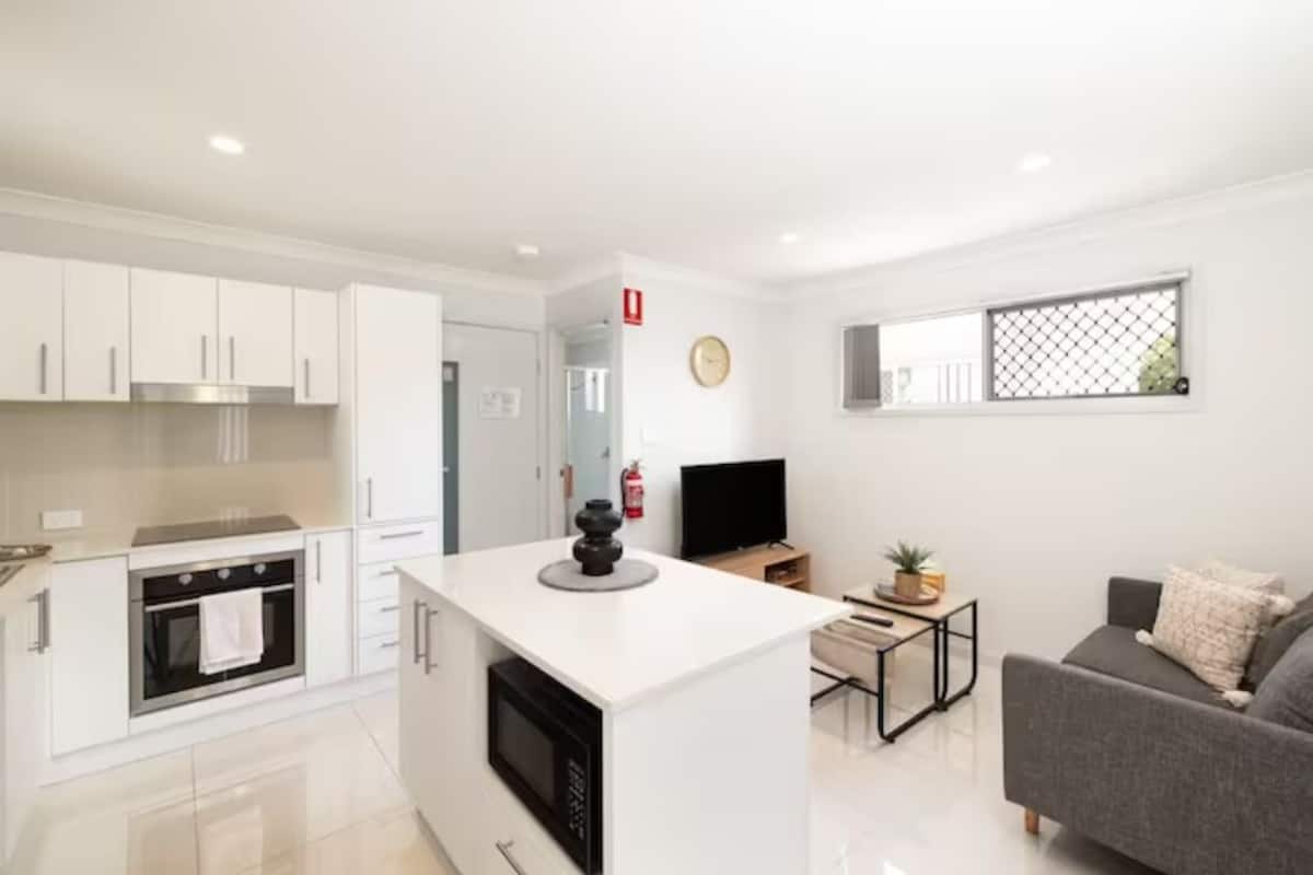 A modern kitchen with white cabinets, a built-in oven, and a microwave. There's a small island with a coffee machine, a TV, and a window with a view.