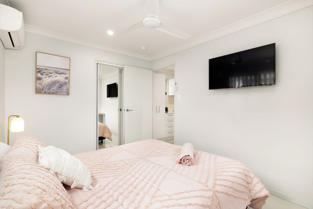 A bedroom with a bed, a ceiling fan, a TV, and a door leading to a bathroom.