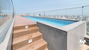 A rooftop pool with wooden steps leading to it, surrounded by a glass railing and a cityscape view. - Loft in Barranco | Sea View (Barranco)