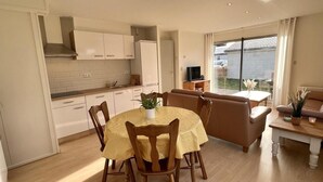 Una cocina moderna con armarios blancos, una mesa de comedor con sillas y un sofá en una sala de estar luminosa con un ventanal grande.
