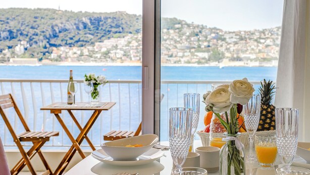 A dining table set with a bowl, glasses, and a bottle, overlooking a coastal view with buildings and hills. - Le Lido, Spectacular Beachfront Cap Ferrat  (Saint-Jean-Cap-Ferrat)