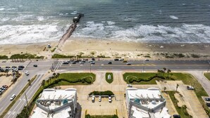 Vue aérienne d’une zone côtière comprenant une plage, une jetée et un parking bordé de plusieurs bâtiments.