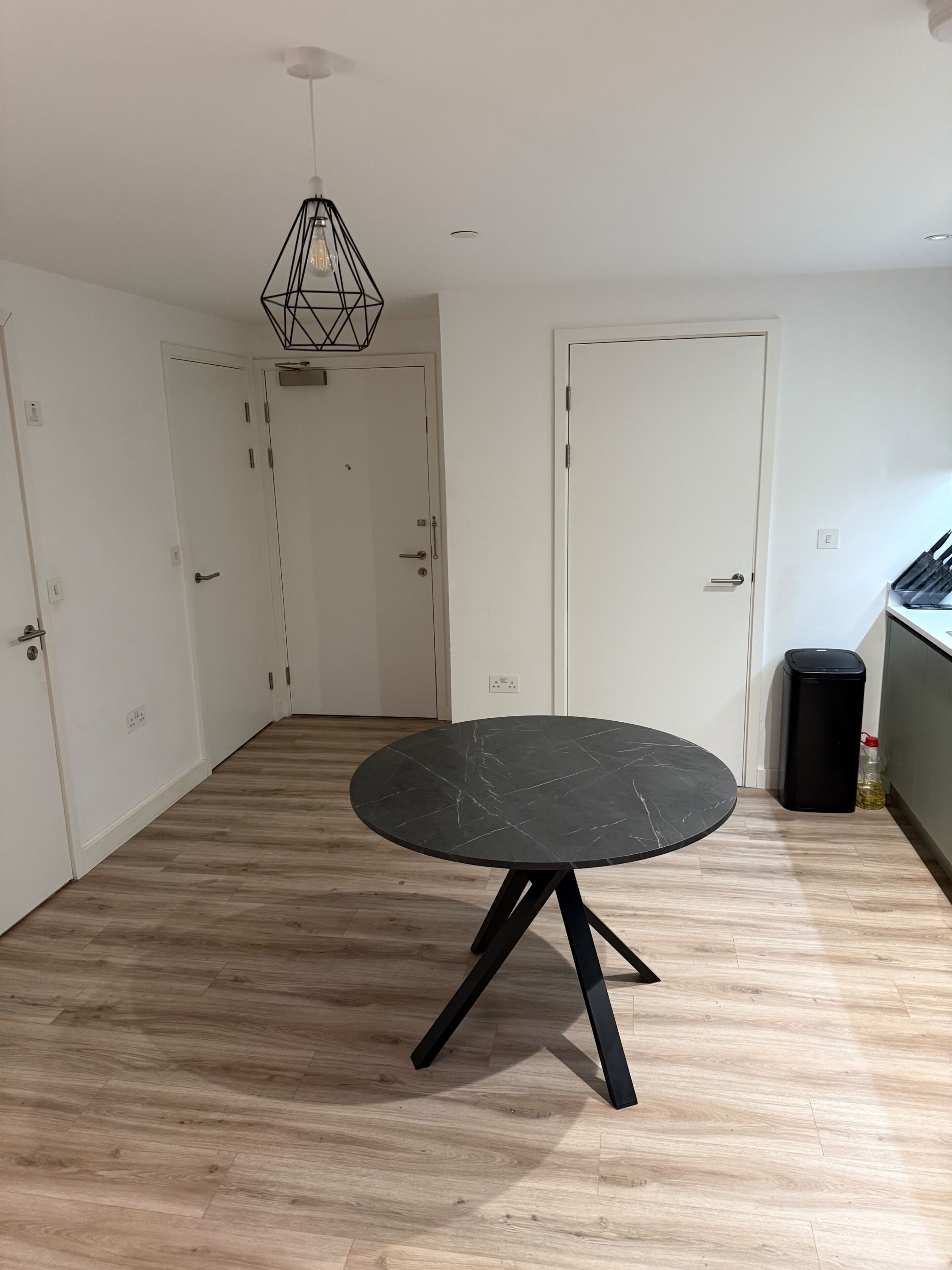 A room with a round table, wooden flooring, white walls, and a black trash bin.