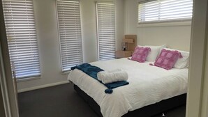 Quarto com cama, paredes brancas e janelas com persianas.