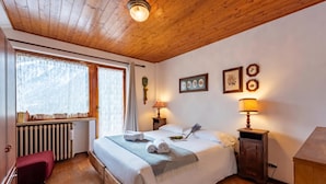 A bedroom with a wooden ceiling, a bed with white linens, two bedside lamps, a radiator, and a window with curtains. - Residence Dolonne, Courmayeur, Italy (Courmayeur)