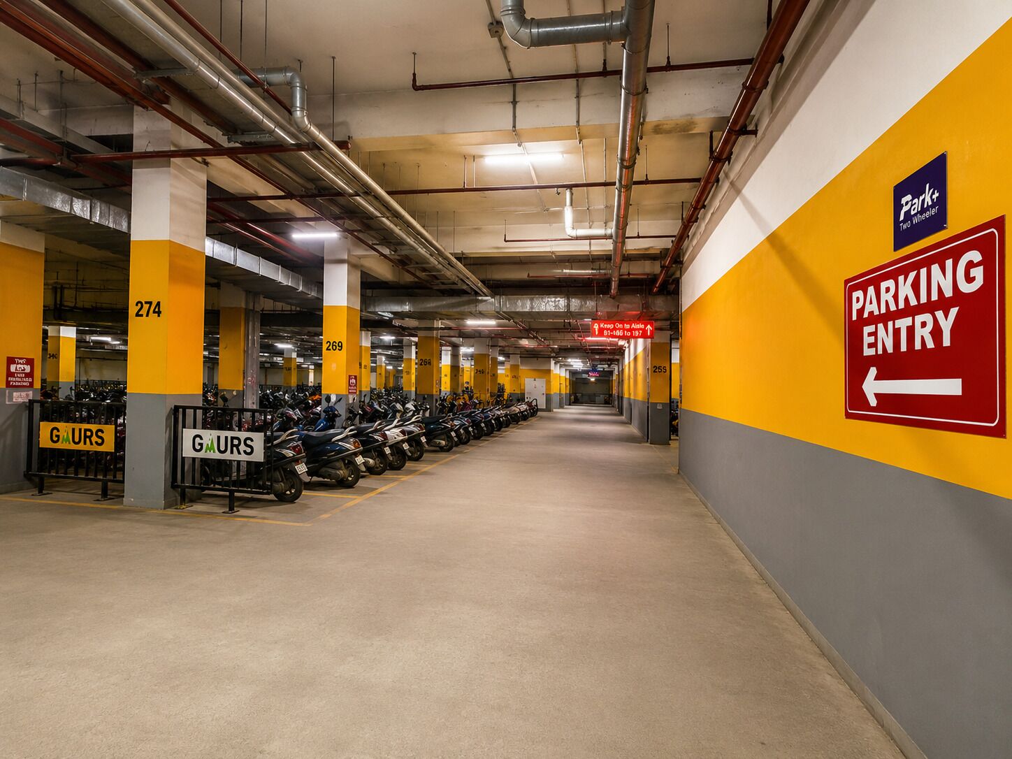A parking garage with rows of parked motorcycles, a 'Parking Entry' sign, and a 'Park+ Two Wheeler' sign.