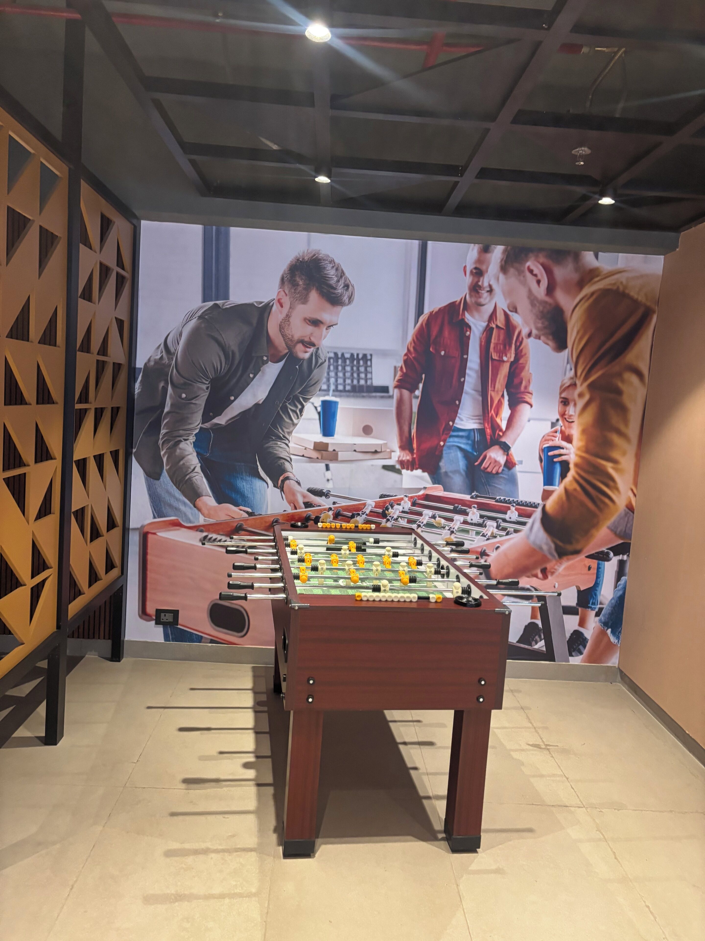 A foosball table in a room with a geometric wall design and a ceiling with exposed beams and lighting fixtures.