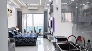 A modern hotel room with a large bed, a sofa, a kitchen area with a sink, and a view of the city. - The Cosette Corner @Gaur city center (Greater Noida)